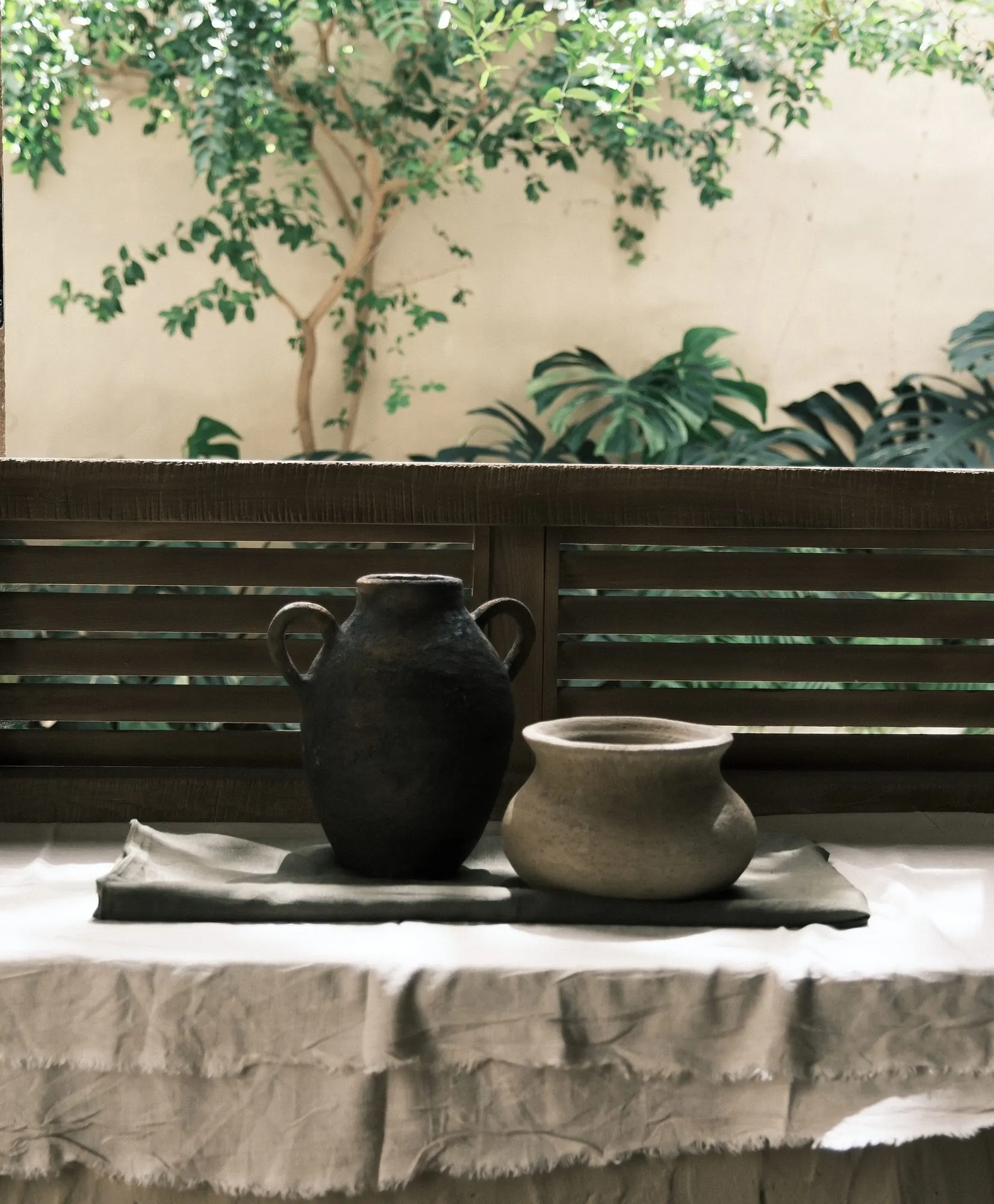 Hũ sẫm màu và nồi thấp màu nhạt trên bàn phủ vải lanh dưới tán cây
